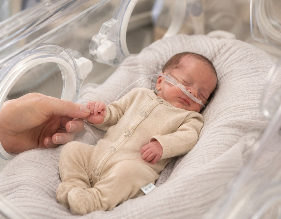 Premature baby resting in a NICU isolette wearing a soft fawn-colored snap-front onesie while holding a parent's finger