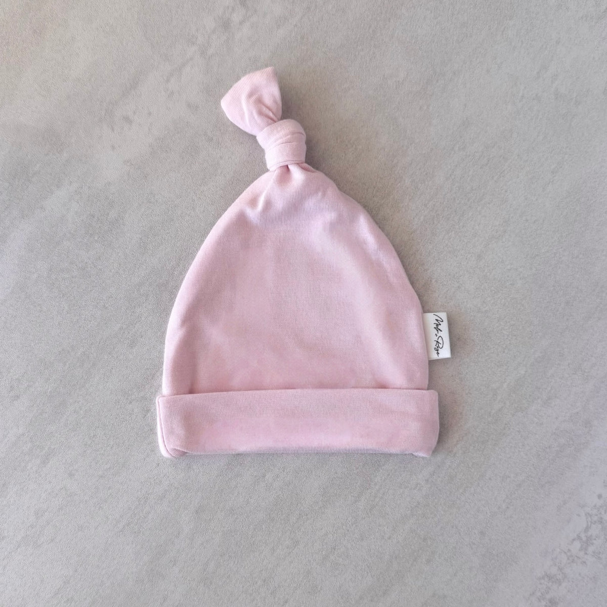 Pink tiny baby hat with a knot on top, featuring a label, on a light gray background