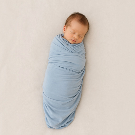 A sleeping premature baby swaddled in a soft light blue wrap, lying on a neutral beige fabric background.