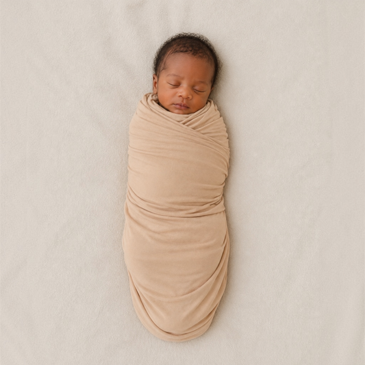 A sleeping premature baby swaddled in a soft, draped tan fabric wrap, lying on a textured light beige blanket.